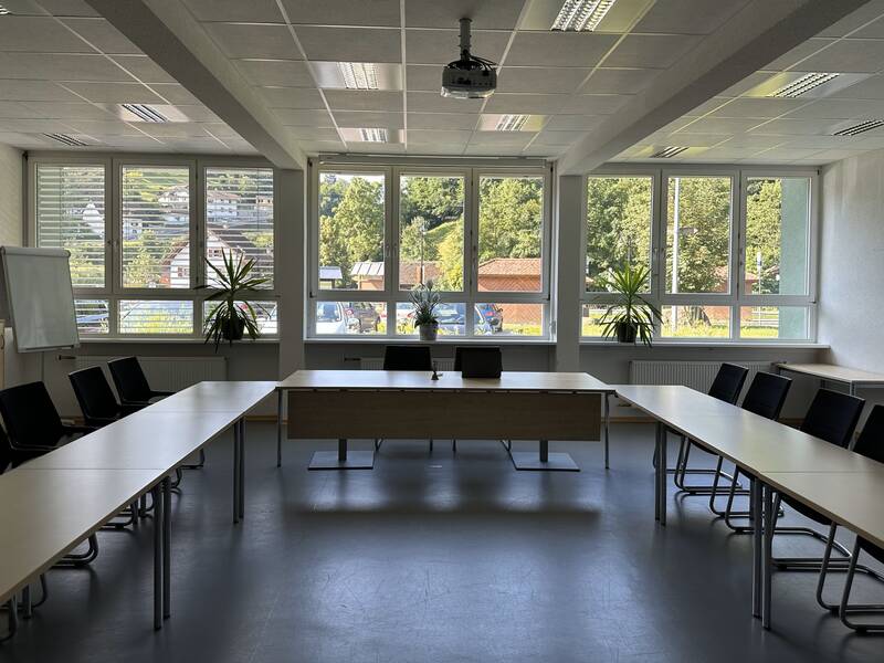 Ein Konferenzraum mit großen Fenstern, rechteckigen Tischen in U-Form, schwarzen Stühlen und Pflanzen im Hintergrund.