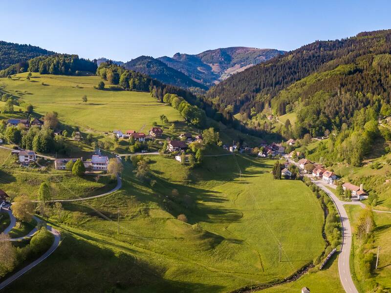 Eine idyllische, hügelige Landschaft mit grünen Wiesen, vereinzelten Häusern und dicht bewaldeten Berghängen im Hintergrund unter einem klaren blauen Himmel.