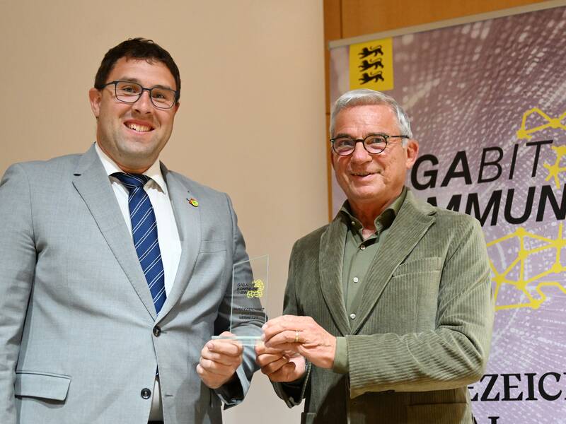 Zwei Männer stehen nebeneinander, während der ältere eine Glas-Trophäe hält; im Hintergrund ist ein Plakat mit der Aufschrift "GIGABIT KOMMUNE" zu sehen.