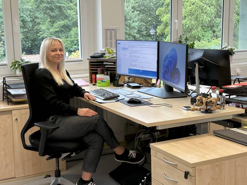 Eine Frau mit blonden Haaren sitzt an einem Schreibtisch mit zwei Computerbildschirmen in einem Büro und schaut lächelnd in die Kamera.
