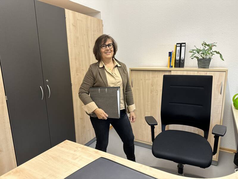 Eine Frau steht lächelnd in einem modernen Büro neben einem schwarzen Bürostuhl, hält einen Ordner und ist von Holzmöbeln umgeben.