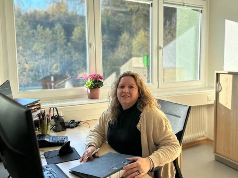 Eine Person sitzt an einem Schreibtisch in einem Büro mit großen Fenstern, durch die man Bäume sieht.