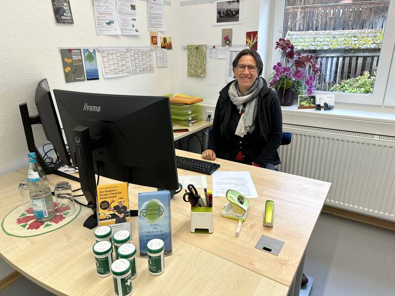 Eine Person sitzt lächelnd an einem Schreibtisch mit Computerbildschirm, Büromaterial und dekorativer Pflanze im Hintergrund.