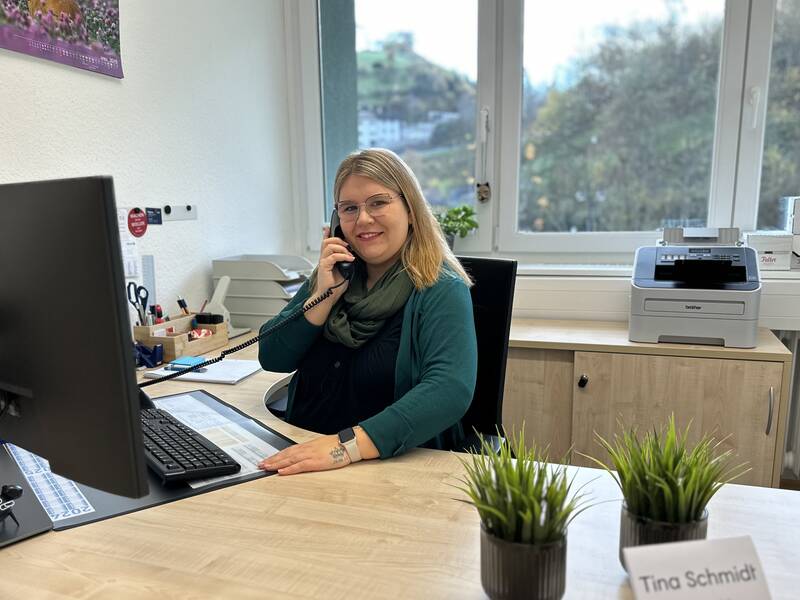 Eine Frau sitzt lächelnd an einem Schreibtisch, telefoniert und hat einen Computerbildschirm vor sich.