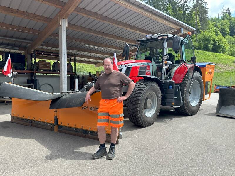 Ein Mann in Arbeitskleidung steht vor einem großen roten Traktor mit Schneepflug, unter einem überdachten Unterstand.