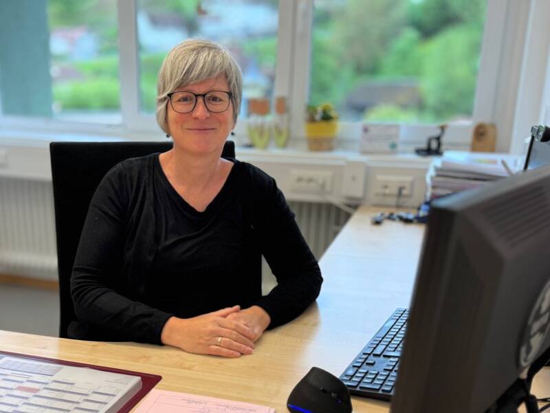 Eine Frau mit grauen Haaren und Brille sitzt lächelnd an einem Schreibtisch mit Computer in einem Büro mit Blick auf Bäume durch das Fenster.